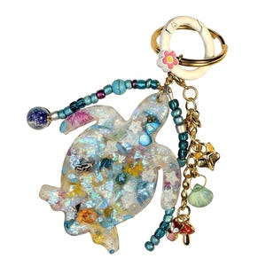 May include: A whimsical keychain featuring a turtle-shaped charm filled with iridescent glitter and small embellishments. The keychain includes a gold-tone ring, a beaded strand, and dangling charms like stars, a seashell, and a mushroom. The turtle charm is approximately 3 inches long.