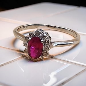 May include: A silver ring featuring a vibrant oval-cut ruby gemstone, surrounded by a halo of smaller, clear gemstones. The ring's band is slender and polished, reflecting light. The setting highlights the ruby's rich color.