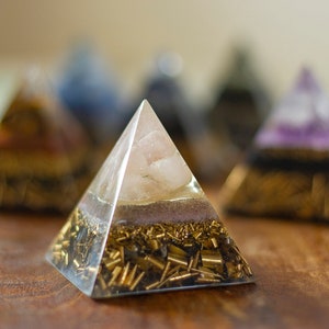 May include: A clear crystal pyramid with a pink rose quartz crystal inside. The pyramid is layered with gold flakes and black stones.