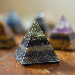 May include: A set of five small crystal pyramids, each with a different color and pattern. The pyramids are made of clear resin and contain various crystals, including amethyst, pyrite, and black tourmaline. The pyramids are arranged on a wooden surface.
