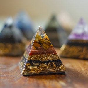 May include: A clear crystal pyramid with layers of different colored stones, including gold, red, and black. The pyramid is sitting on a wooden surface.