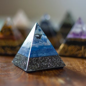 May include: A blue, white, and black pyramid with a cloudy sky design. The pyramid is made of resin and is sitting on a wooden surface.