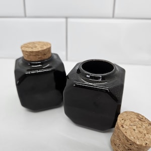 May include: Two black, octagonal glass jars with cork stoppers. One jar has a cork stopper in place, the other is open. A third cork stopper rests beside the open jar. The jars are on a white surface.