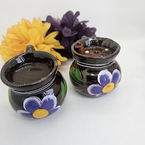 May include: Two small, dark brown ceramic mugs with a floral design. The mugs have a dark liquid inside, and one has a cinnamon stick. Yellow and purple flowers are in the background. The mugs are likely for beverages.