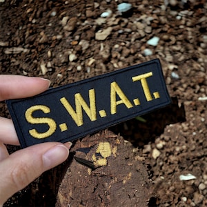 Può includere: Una toppa nera con lettere ricamate in oro che compongono la scritta "S.W.A.T."