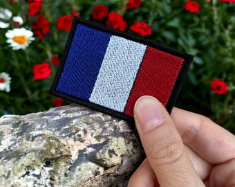 May include: A close-up of a blue, white, and red embroidered patch of the French flag. The patch is being held by a hand against a background of red flowers.