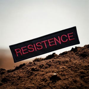 May include: A black rectangular patch with the word "RESISTENCE" embroidered in red thread. The patch is set against a backdrop of brown soil and a neutral background.
