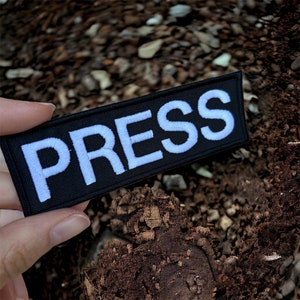 Peut inclure: Un patch en tissu noir avec du texte brodé en blanc qui dit "PRESS".