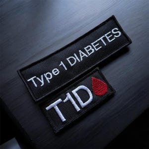 Op de afbeelding: Twee rechthoekige zwarte geborduurde patches. De grotere patch bevat de witte tekst Type 1 DIABETES. De kleinere patch bevat de witte tekst T1D gevolgd door een rode geborduurde druppelvorm, die het symbool van een bloeddruppel voorstelt.