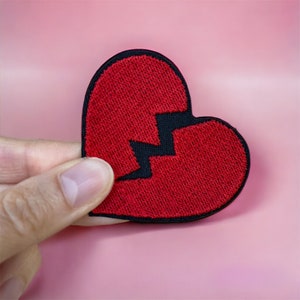 Puede incluir: Parche bordado con forma de corazón roto. El parche es rojo con un borde negro y una línea negra en zigzag que divide el corazón en dos. El parche se sostiene sobre un fondo rosa.