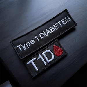 Puede incluir: Dos parches negros bordados. El parche más grande dice "Type 1 DIABETES" en hilo blanco. El parche más pequeño tiene "T1D" en blanco y una gota de sangre roja. Los parches están sobre una superficie oscura.
