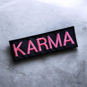 Puede incluir: Parche rectangular negro con la palabra "KARMA" bordada en rosa. El parche tiene un borde negro y está sobre un fondo gris. Las letras están en una fuente en negrita sin serifa.