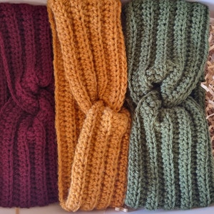 May include: Three handmade crochet headbands in a box. The headbands are in shades of burgundy, mustard yellow, and olive green. Each headband features a twisted knot design and a ribbed texture, showcasing the detailed craftsmanship.
