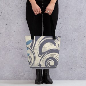 May include: A tote bag featuring a swirling abstract design in shades of blue, grey, and white. The bag has black handles.