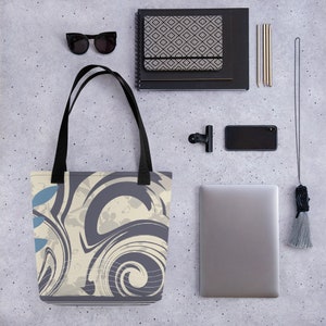 May include: A tote bag with a black handle and a cream and grey abstract swirl design. The bag is on a grey textured surface with other items such as a laptop, sunglasses, and a notebook.