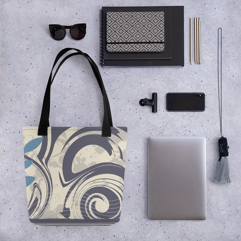 May include: A tote bag with a black handle and a cream and grey abstract swirl design. The bag is on a grey textured surface with other items such as a laptop, sunglasses, and a notebook.