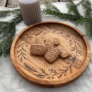 Peut inclure: Un plateau de service en bois avec un motif floral et plusieurs biscuits. Une bougie pilier grise et des brins de conifères sont également présents. Le plateau est en bois brun clair et les biscuits sont de couleur brun clair.