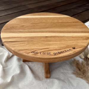 Può includere: Un alzatina rotonda in legno marrone chiaro con un'unica gamba. L'alzatina ha una finitura a venature naturali e le parole "mit LIEBE gebacken" sono incise sul bordo, insieme a un piccolo simbolo a forma di cuore. L'alzatina misura circa 25 cm di diametro.