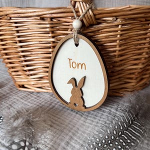 Puede incluir: Una etiqueta de madera blanca en forma de huevo de Pascua con el nombre "Tom" escrito en ella. La etiqueta tiene un recorte de un conejo y está unida a una cesta de mimbre con un asa marrón.