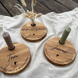 May include: Three wooden candle holders with engraved text and heart details. Each holder has a different name and message, with a candle inserted. The holders are on a white cloth, with dried flowers in one.
