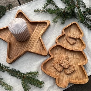 Peut inclure: Plateaux en bois en forme d'étoile et de sapin de Noël. Le plateau en étoile contient une bougie beige. Le plateau en forme d'arbre contient plusieurs petits biscuits. Des branches de pin vertes sont disposées autour des plateaux.