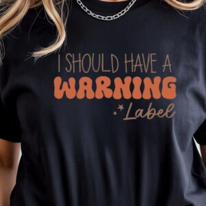 Puede incluir: Camiseta negra con el texto "I SHOULD HAVE A WARNING LABEL" en naranja y marrón, con "*Label" en una fuente cursiva más pequeña. La camiseta es de estilo casual.