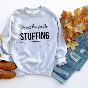 以下が含まれることがあります： グレーのトレーナーに「I'm just here for the STUFFING」という文字がプリントされています。トレーナーは白い背景に置かれ、茶色の靴とダメージ加工が施された青いジーンズが一緒に置かれています。