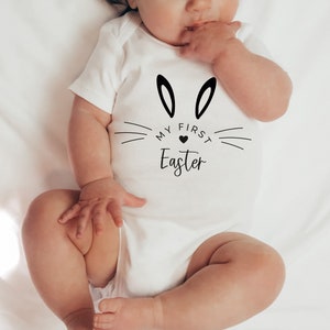 Könnte beinhalten: Weißer Baby-Strampler mit schwarzen Hasenohren und dem Text "Mein erstes Ostern" mit einem kleinen schwarzen Herz.