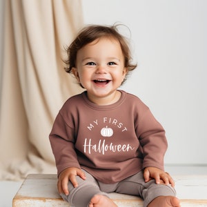 May include: A baby wearing a brown sweatshirt with the text "My First Halloween" and a white pumpkin graphic. The baby is sitting on a white wooden stool and smiling.