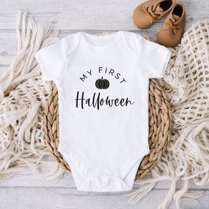 Może przedstawiać: Biały śpioszek dla niemowlęcia z czarnym napisem "Moje pierwsze Halloween" i czarną grafiką dyni.