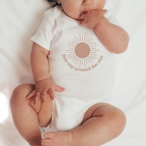 May include: A white baby onesie with a brown sun graphic and the text "first trip around the sun".