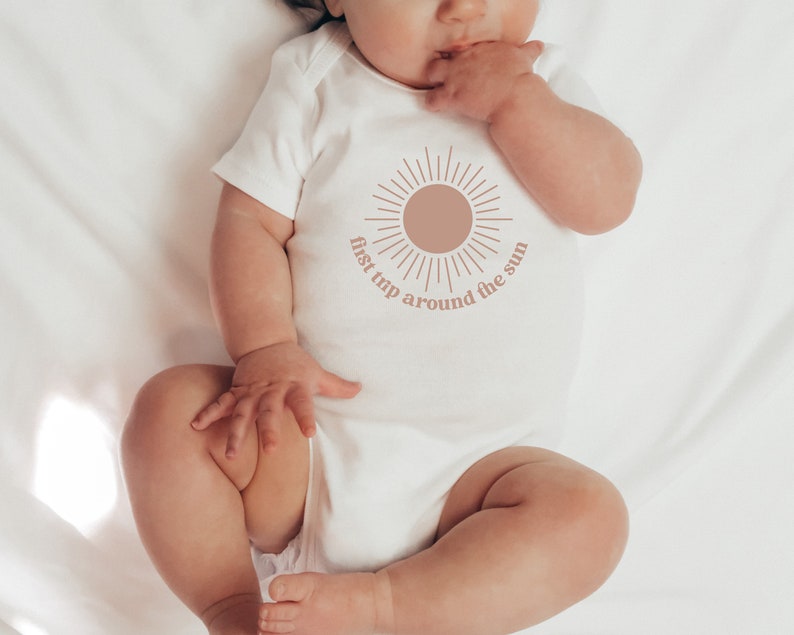 May include: A white baby onesie with a brown sun graphic and the text "first trip around the sun".