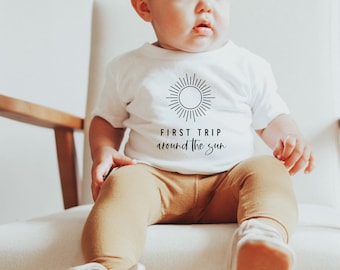 May include: A white baby t-shirt with a black sun graphic and the text "FIRST TRIP around the sun." The baby is wearing tan pants and sitting in a chair. The shirt is made of soft material.