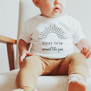 May include: A white baby onesie with a black graphic of a sun and the text "first trip around the sun".