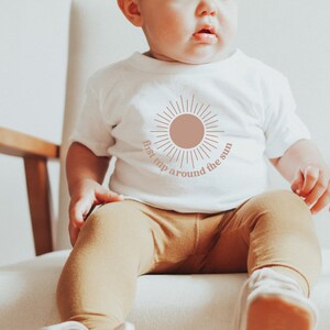 May include: A white baby onesie with a pink sun graphic and the text "first trip around the sun". A baby is wearing the onesie and sitting in a wooden chair.