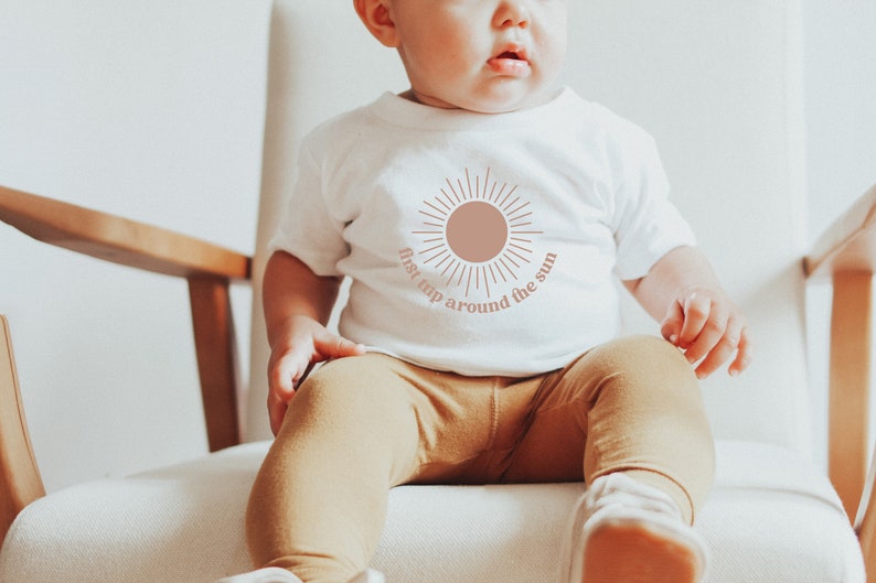 May include: A white baby onesie with a pink sun graphic and the text "first trip around the sun". A baby is wearing the onesie and sitting in a wooden chair.