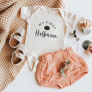 May include: A white baby onesie with the text "My First Halloween" and a black pumpkin graphic. The onesie is paired with a pair of light pink baby bloomers and a pair of white baby shoes.
