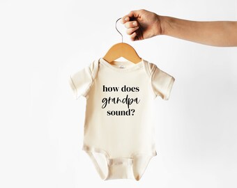 May include: Cream-colored baby onesie with the text "how does grandpa sound?" in black script. The onesie is hanging on a wooden hanger, held by a hand against a white background. A cute and simple design for a baby.