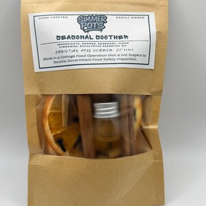 May include: A brown paper bag with a clear window, containing dried orange slices, cinnamon sticks, and a small bottle. The label reads "Seasonal Soother" with ingredients listed. The bag is labeled "Hand Crafted" and "Family Owned."
