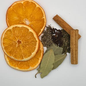 May include: Overhead shot of dried orange slices, cinnamon sticks, bay leaves, cloves, and dried herbs. The ingredients are arranged on a white surface, suggesting a culinary or aromatherapy theme. The orange slices are a vibrant orange color.
