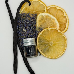 May include: An arrangement of dried lemon slices, lavender buds, and vanilla beans. A small glass bottle with a silver lid contains a dark liquid. The items are arranged on a white surface, creating a natural, aromatic display.