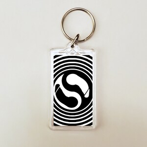 May include: A clear rectangular keychain with a silver metal ring. The keychain features a black and white graphic design with a swirling pattern and a white shape in the center. The design is encased in a clear plastic holder.