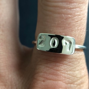Peut inclure: Une bague en argent avec une face rectangulaire gravée du chiffre "2017". La bague a un design simple, avec un fin anneau et une surface polie et réfléchissante. Les chiffres sont clairement gravés dans le métal.