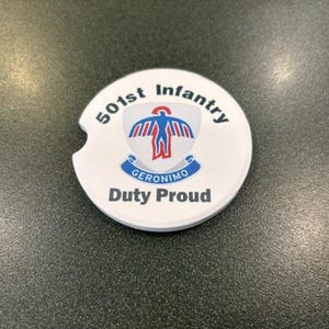May include: A round white ceramic car coaster featuring a small notch on the left side. The design includes the text 501st Infantry at the top, a central shield emblem with a blue and red bird graphic, the word Geronimo on a blue banner, and Duty Proud.