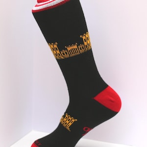 May include: Black sock with a red heel and toe, and a red, white, and black striped cuff. The sock features a gold-colored architectural design near the top and the word "BER" in gold near the ankle.
