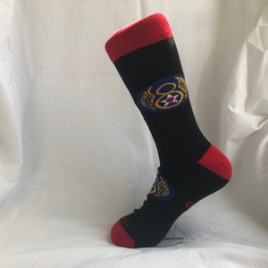 Puede incluir: Calcetines negros con un puño rojo y un emblema azul y dorado con el número 8 y alas. El emblema está en la parte delantera del calcetín.