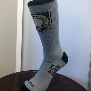 May include: A pair of gray crew socks with a black and white graphic of a military insignia on the top and the words "Duty Proud" on the bottom.