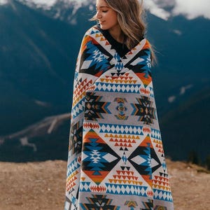 May include: A large rectangular blanket featuring a repeating geometric pattern in shades of orange, blue, black, and white. The design consists of stacked triangles, diamonds, and linear motifs arranged in horizontal bands across a light gray background.
