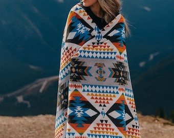 May include: A large rectangular blanket featuring a repeating geometric pattern in shades of orange, blue, black, and white. The design consists of stacked triangles, diamonds, and linear motifs arranged in horizontal bands across a light gray background.