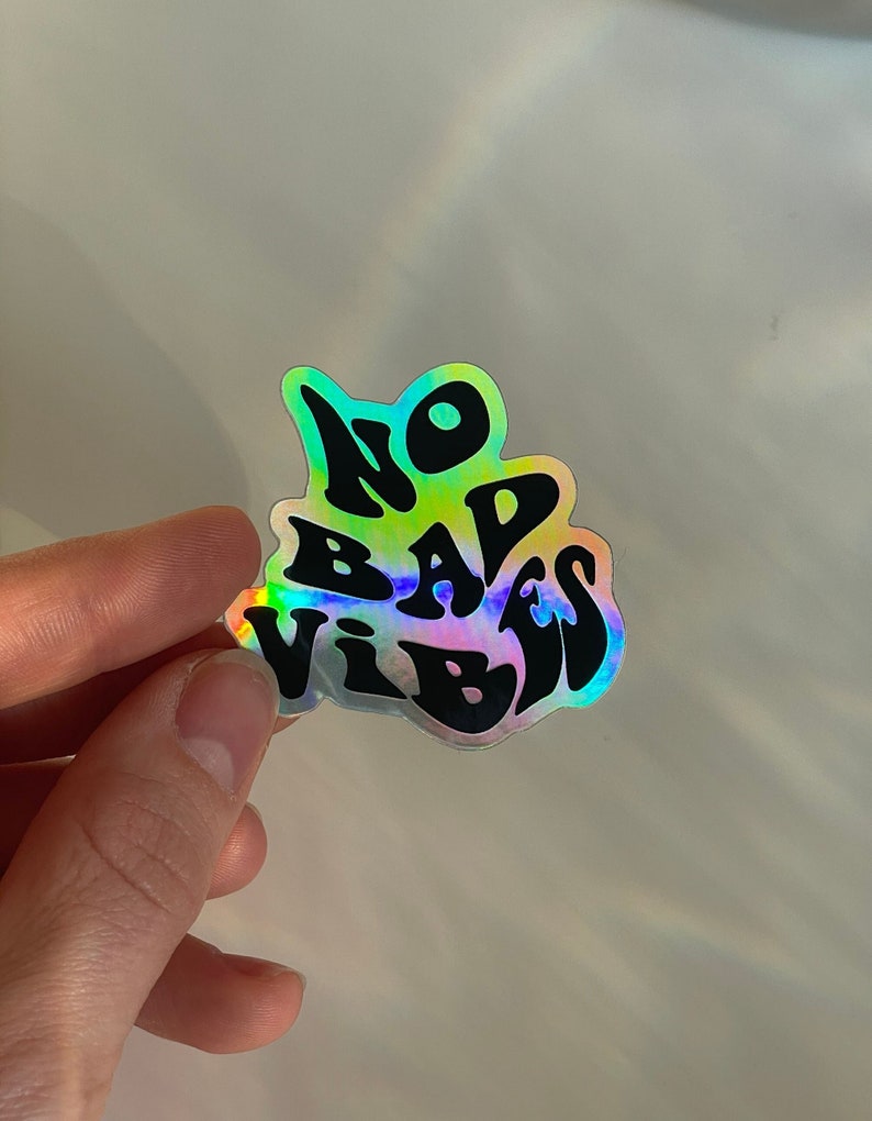 以下が含まれることがあります： 「NO BAD VIBES」の文字が黒色のスタイリッシュな書体で書かれたホログラフィックステッカー。ステッカーは虹色のイリデッセント効果があり、表面全体で色が変化します。無地の明るい背景にステッカーが貼られています。